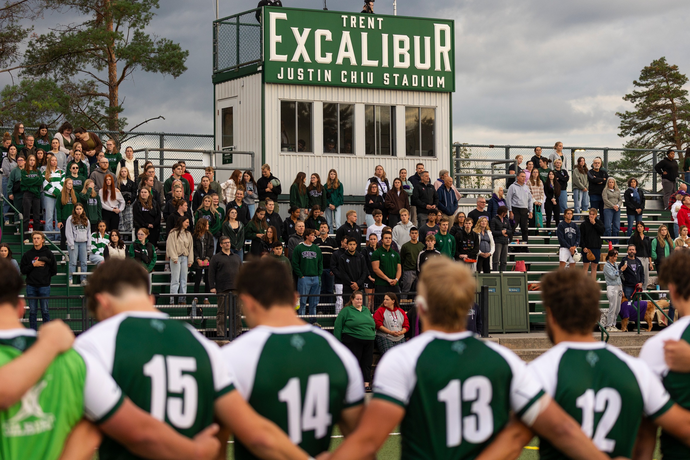 Six Excalibur men's rugby players stand with their arms linked, facing a full stadium of fans.